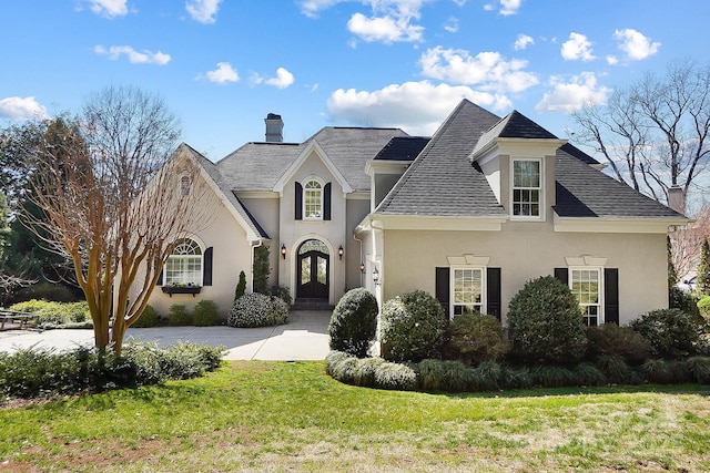 french provincial home featuring a front yard, stucco siding, french doors, a chimney, and driveway