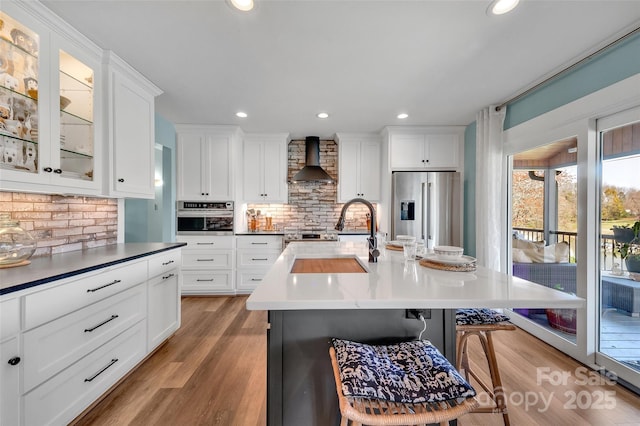 kitchen featuring light wood-type flooring, appliances with stainless steel finishes, a breakfast bar area, white cabinets, and wall chimney range hood