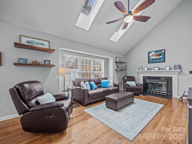 living room featuring a glass covered fireplace, wood finished floors, a skylight, baseboards, and ceiling fan