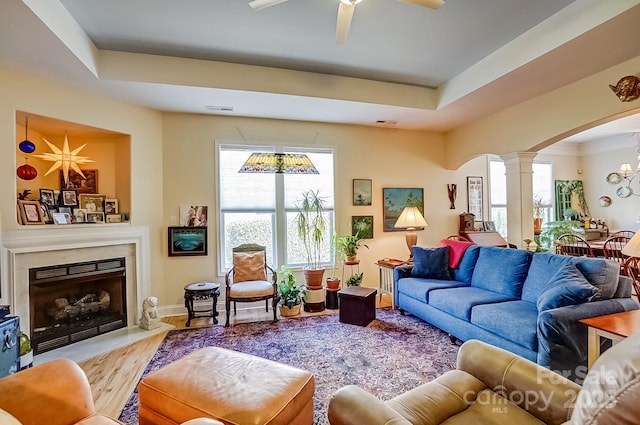 living area with ceiling fan, a tray ceiling, a premium fireplace, decorative columns, and arched walkways