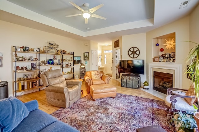 living area featuring visible vents, a tray ceiling, wood finished floors, a high end fireplace, and ceiling fan