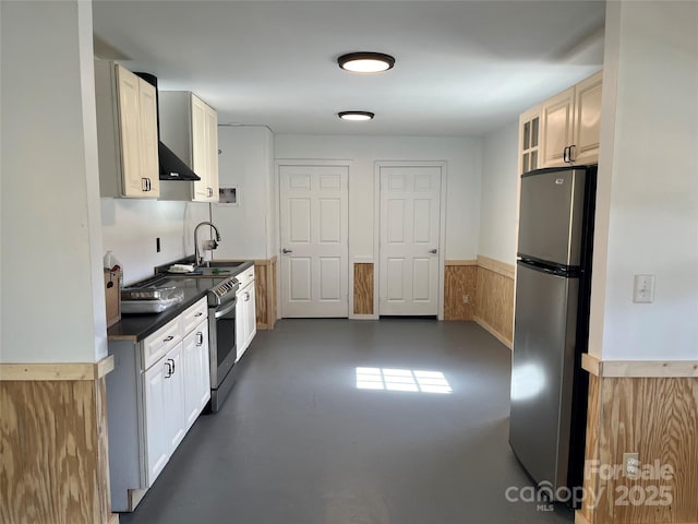 kitchen with a sink, concrete flooring, appliances with stainless steel finishes, dark countertops, and wall chimney range hood