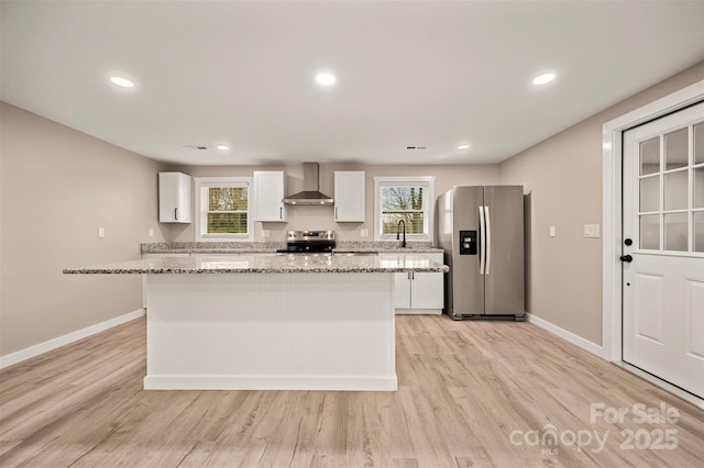 kitchen with light wood finished floors, a kitchen island, white cabinets, appliances with stainless steel finishes, and wall chimney range hood