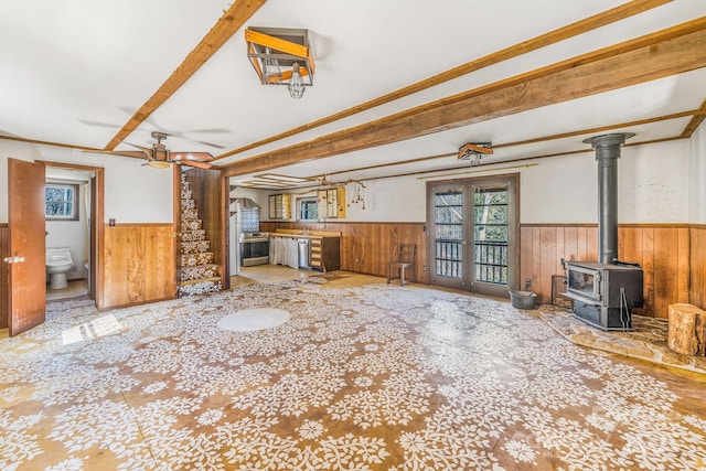 unfurnished living room with wooden walls, a ceiling fan, a wood stove, wainscoting, and beamed ceiling