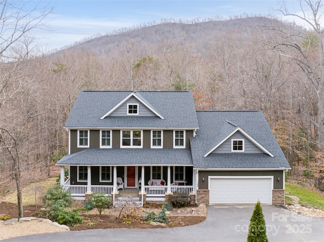 view of front facade featuring a wooded view, roof with shingles, covered porch, concrete driveway, and stone siding