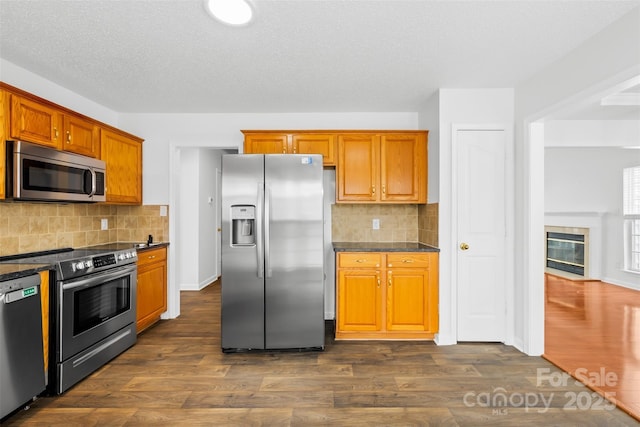 kitchen featuring backsplash, a tiled fireplace, brown cabinets, stainless steel appliances, and dark wood-style flooring