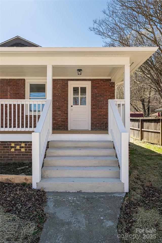 view of exterior entry featuring brick siding, covered porch, and fence