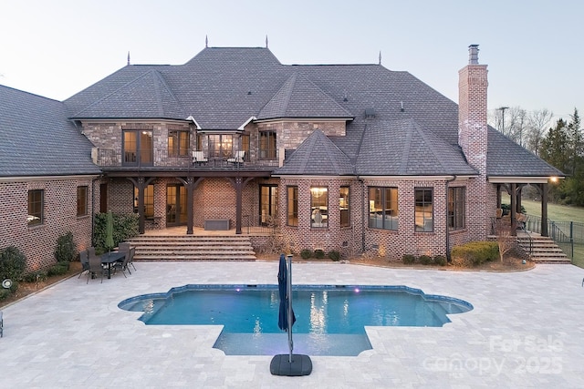 rear view of property featuring a patio, a balcony, an outdoor pool, a chimney, and stone siding