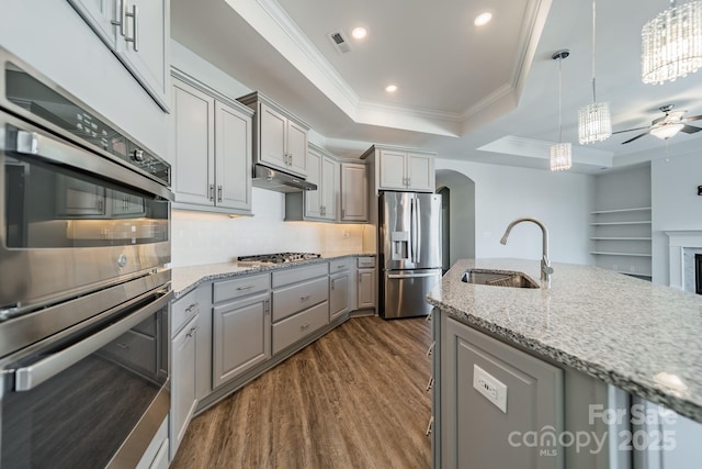 kitchen with gray cabinetry, under cabinet range hood, appliances with stainless steel finishes, arched walkways, and a sink