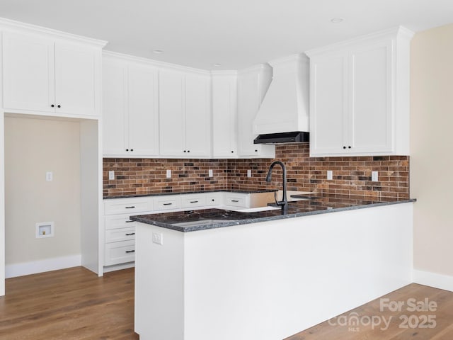 kitchen with backsplash, custom range hood, dark stone countertops, wood finished floors, and white cabinetry