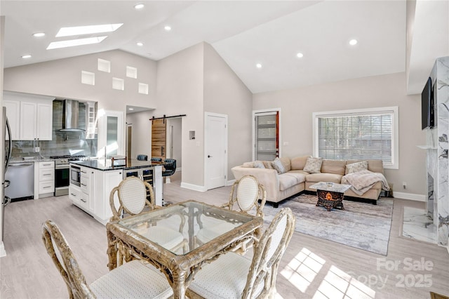 dining area featuring high vaulted ceiling, light wood-style flooring, a barn door, a skylight, and baseboards