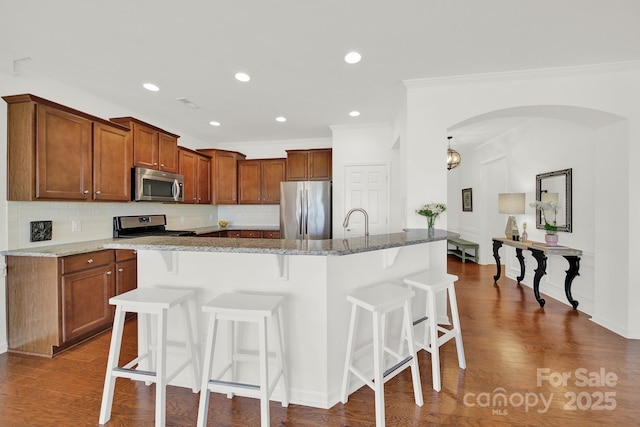 kitchen featuring brown cabinetry, stone counters, arched walkways, appliances with stainless steel finishes, and a kitchen breakfast bar