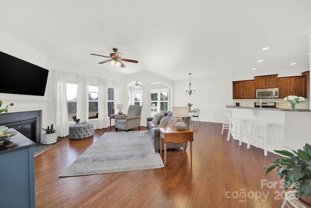 living area featuring dark wood-type flooring, ceiling fan with notable chandelier, a glass covered fireplace, arched walkways, and a decorative wall