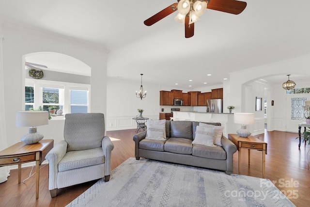 living room featuring wood finished floors, arched walkways, wainscoting, a decorative wall, and ceiling fan with notable chandelier