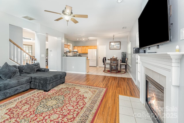 living area featuring visible vents, light wood-style flooring, a ceiling fan, stairway, and a tile fireplace