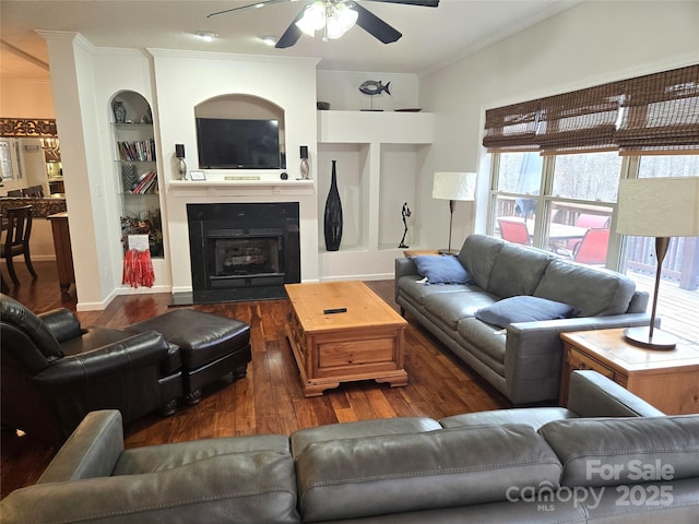 living area with built in shelves, a fireplace with flush hearth, a ceiling fan, wood finished floors, and crown molding