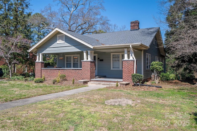 view of front facade with brick siding, a shingled roof, a front lawn, covered porch, and a chimney