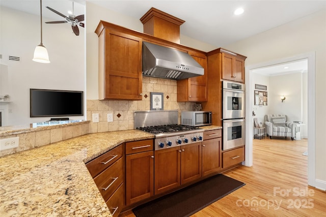 kitchen with brown cabinets, stainless steel appliances, wall chimney exhaust hood, light wood finished floors, and decorative backsplash