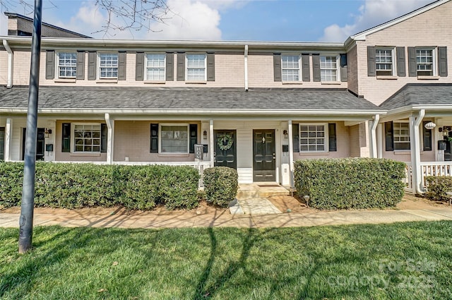 townhome / multi-family property with brick siding, a porch, a front yard, and roof with shingles