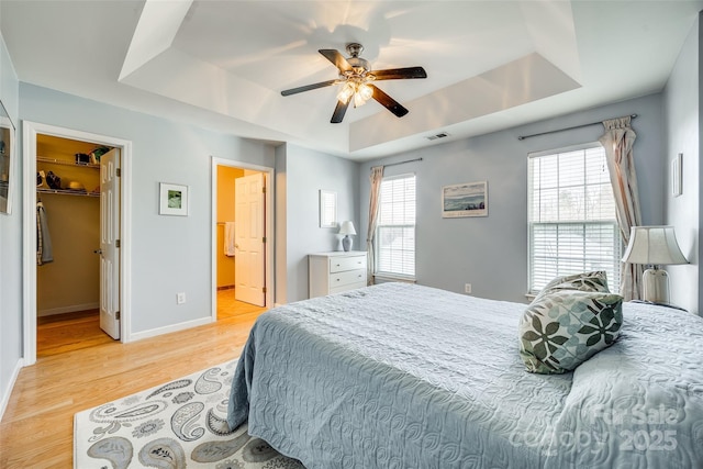 bedroom featuring a walk in closet, a tray ceiling, light wood-style flooring, and baseboards