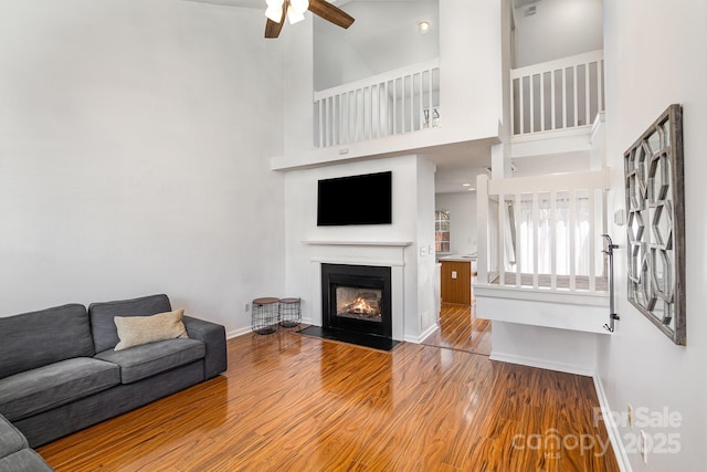 living room featuring a fireplace with flush hearth, a ceiling fan, wood finished floors, a high ceiling, and baseboards