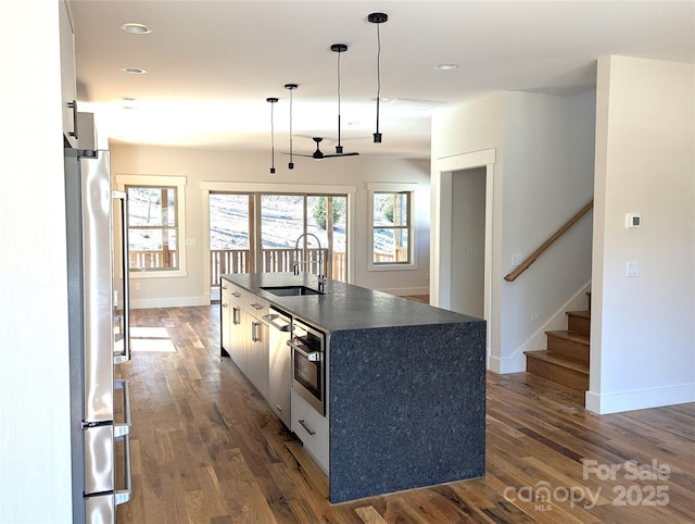 kitchen with dark countertops, dark wood finished floors, a center island with sink, stainless steel appliances, and a sink