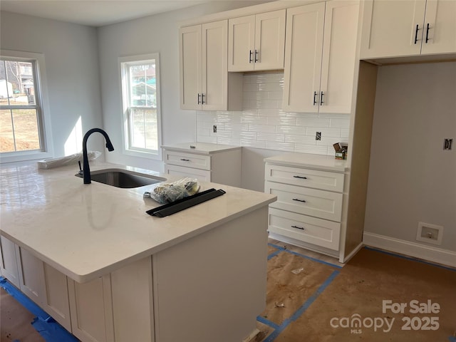 kitchen with an island with sink, a sink, white cabinetry, decorative backsplash, and baseboards