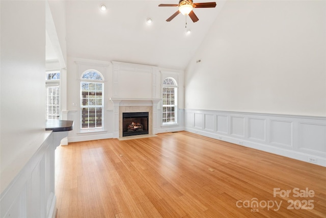 unfurnished living room featuring a ceiling fan, high vaulted ceiling, a fireplace with flush hearth, wainscoting, and light wood-type flooring