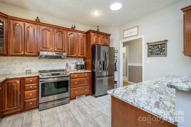 kitchen with under cabinet range hood, light stone counters, backsplash, stainless steel appliances, and glass insert cabinets