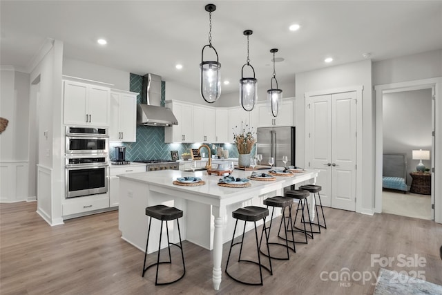 kitchen featuring an island with sink, a kitchen breakfast bar, white cabinetry, stainless steel appliances, and wall chimney range hood