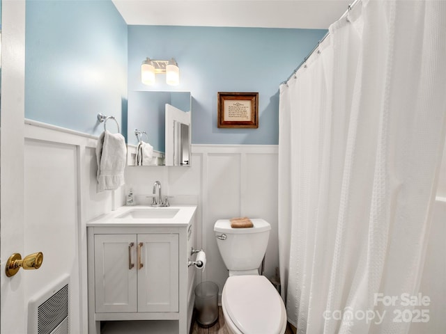 bathroom with a wainscoted wall, toilet, heating unit, a decorative wall, and vanity