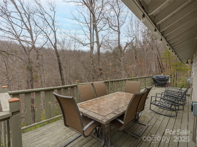 wooden deck with a view of trees, a grill, and outdoor dining space