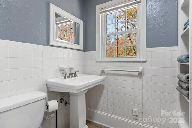 bathroom featuring toilet, a sink, tile walls, wainscoting, and a textured wall