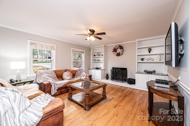 living room featuring light wood-type flooring, ornamental molding, a ceiling fan, built in features, and a brick fireplace