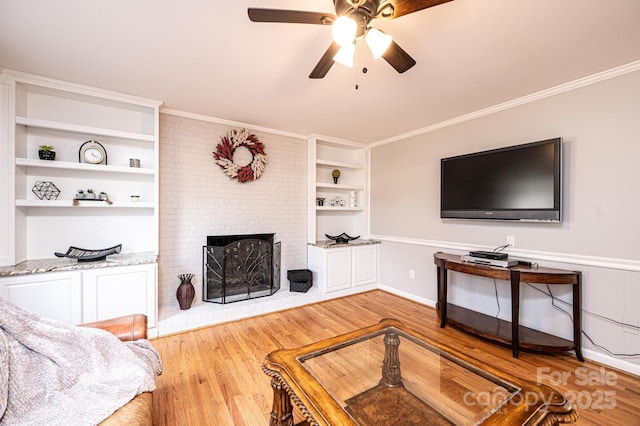 living area with a brick fireplace, built in shelves, crown molding, wood finished floors, and a ceiling fan