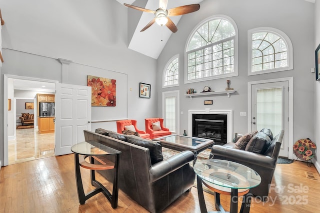 living area featuring visible vents, light wood-style floors, a glass covered fireplace, high vaulted ceiling, and a ceiling fan