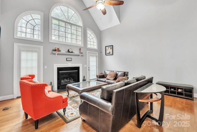 living area featuring a fireplace with flush hearth, high vaulted ceiling, light wood-style flooring, a ceiling fan, and baseboards