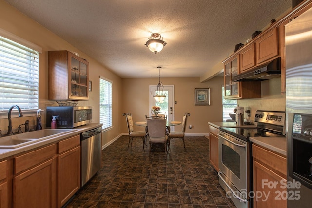 kitchen with under cabinet range hood, a sink, appliances with stainless steel finishes, brown cabinetry, and glass insert cabinets