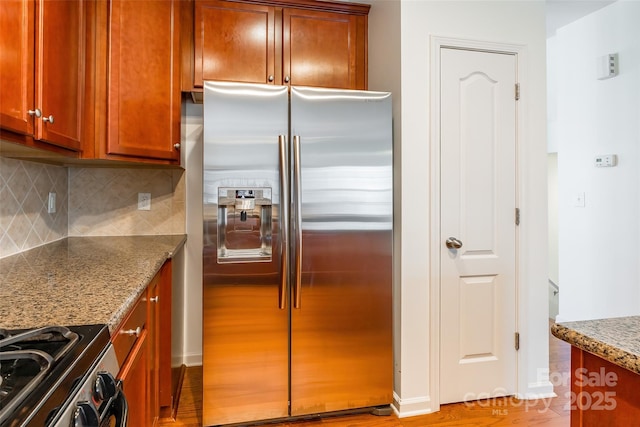 kitchen featuring baseboards, decorative backsplash, stone counters, appliances with stainless steel finishes, and brown cabinets