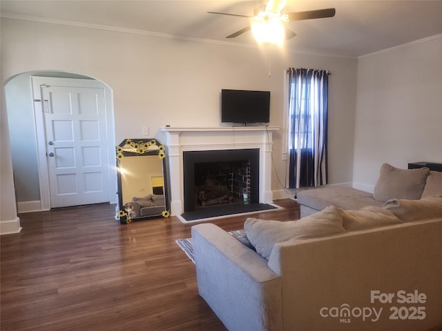 living room with a fireplace with raised hearth, a ceiling fan, wood finished floors, arched walkways, and crown molding