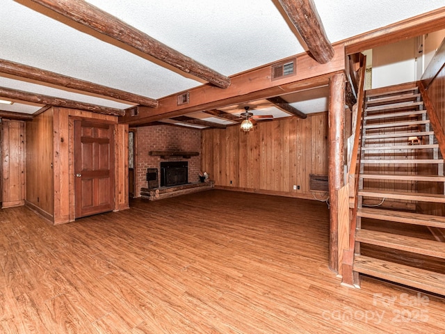 unfurnished living room featuring wooden walls, visible vents, a brick fireplace, stairway, and light wood-style floors