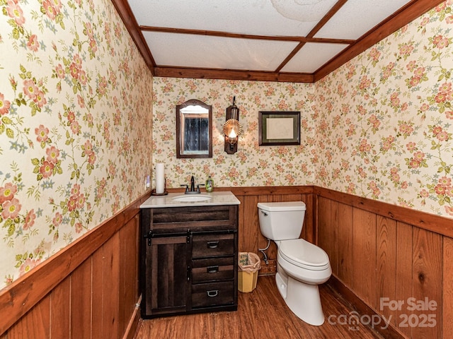 half bath with a wainscoted wall, toilet, wood finished floors, wallpapered walls, and vanity