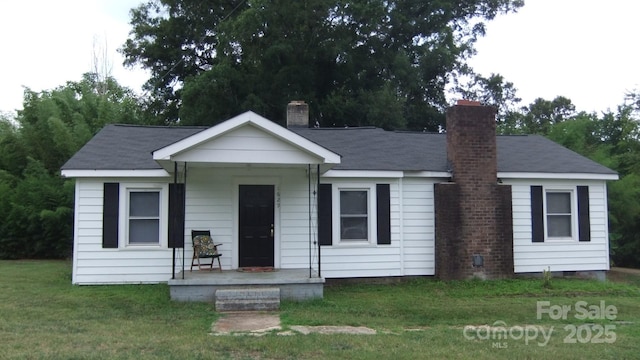 view of front facade with a shingled roof, a front lawn, covered porch, and a chimney