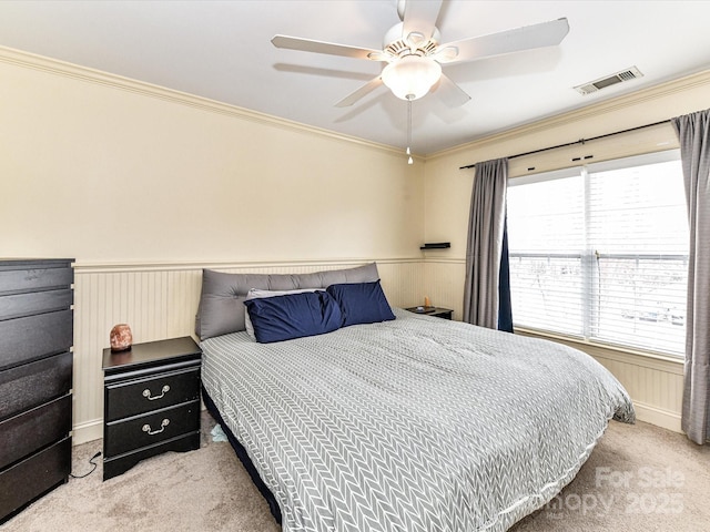 carpeted bedroom with a wainscoted wall, crown molding, visible vents, and ceiling fan