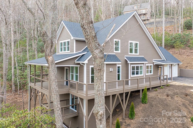 rear view of house featuring a garage, a view of trees, driveway, and a shingled roof