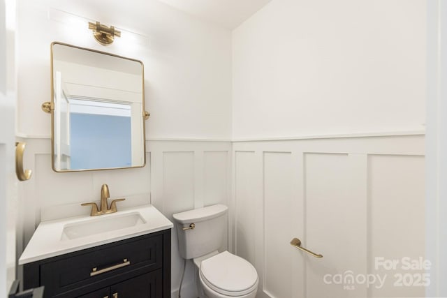 bathroom featuring a decorative wall, wainscoting, toilet, and vanity