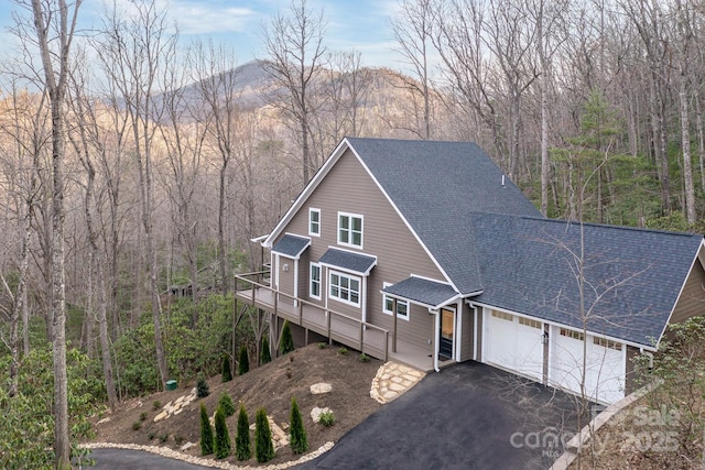 view of front of house with a wooded view, a shingled roof, a garage, a deck, and aphalt driveway