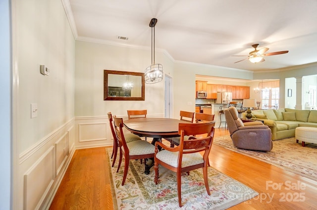 dining room with visible vents, a wainscoted wall, ornamental molding, ceiling fan with notable chandelier, and light wood finished floors