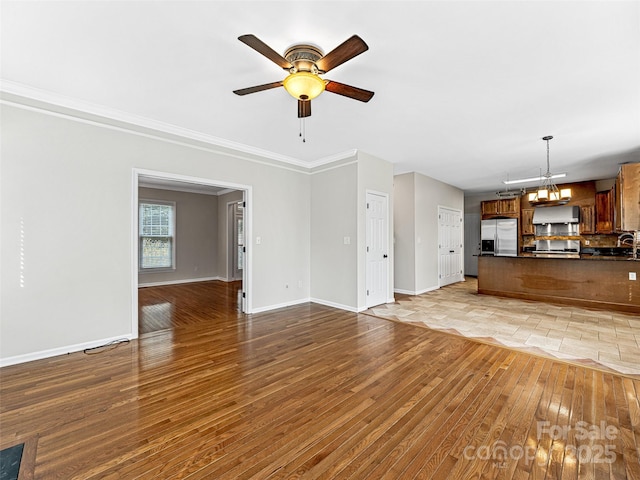 unfurnished living room featuring ornamental molding, ceiling fan with notable chandelier, light wood-style floors, and baseboards