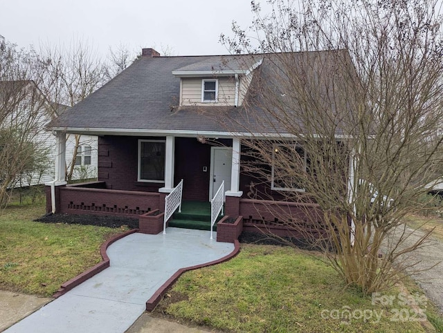 view of front facade with a porch, roof with shingles, a front yard, brick siding, and a chimney
